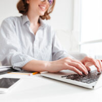 Cropped photo of pretty brunette woman typing email on laptop computer while sitting at home, selective focus on hand